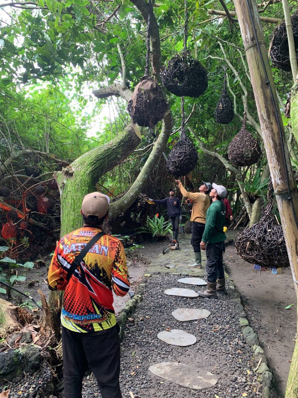 Aurora Park during installation — sculptural woven structures hang above a stone path, a person in a printed jacket walks ahead.