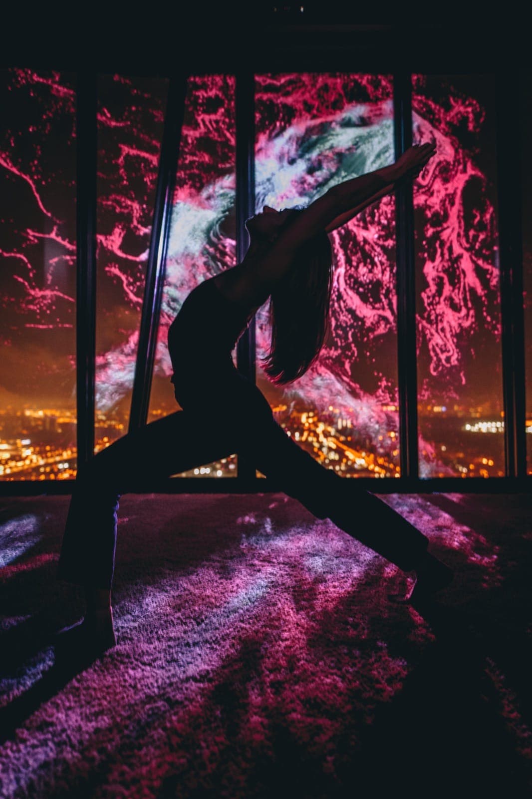 A figure in warrior pose silhouetted against a panoramic window painted with a vivid pink-and-blue cosmic projection over the night city.