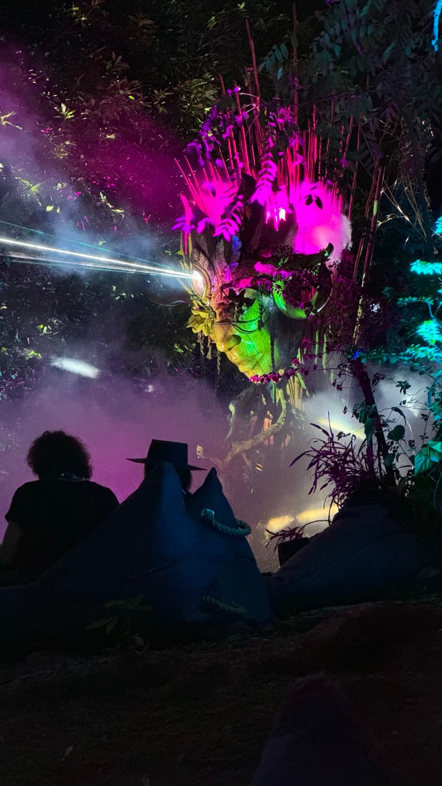 Kriya at night with magenta and green lasers, fog rising, audience seated on bean-bag pyramids in the foreground.