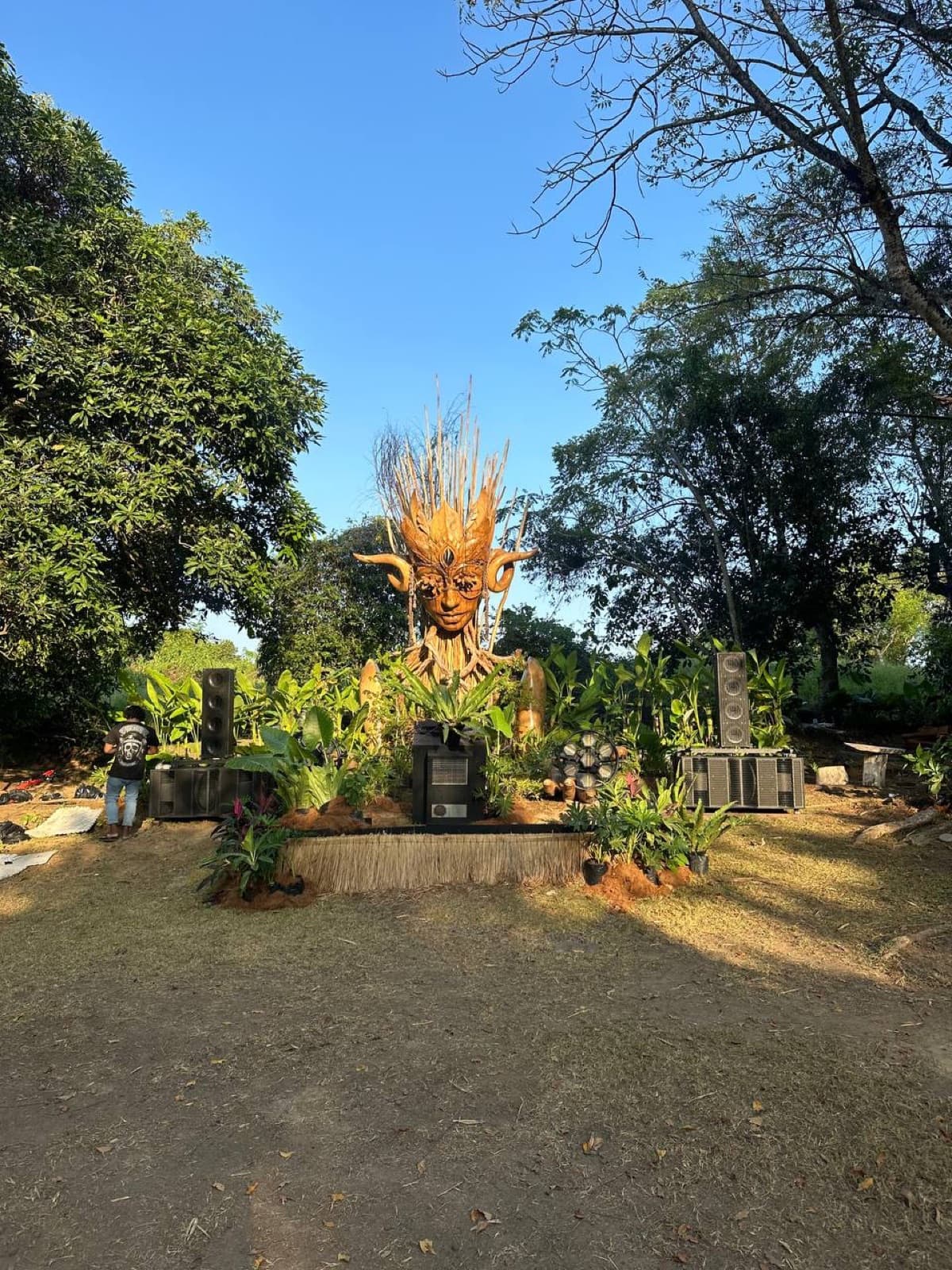 Kriya in daytime — sculpted face crowned with golden grasses, flanked by stacked speakers, set into a clearing at Nuanu.