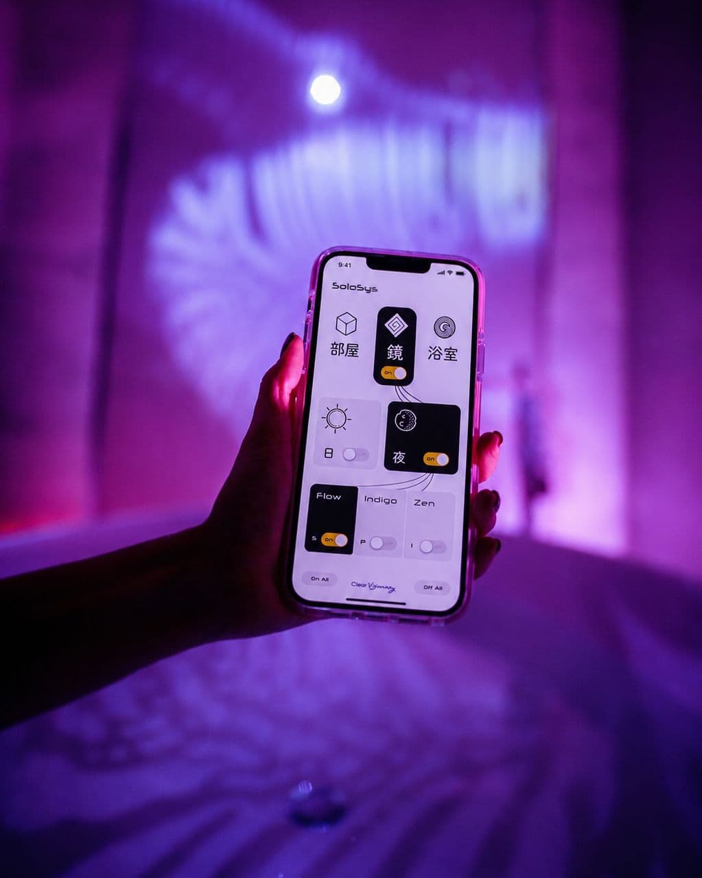 An iPhone held in front of a purple-lit bath, showing the SoloSys control panel — Projections, Interactive Art, Audio-reactive Art, Art Gallery, Infinity Mirror, Bathroom, Sound.