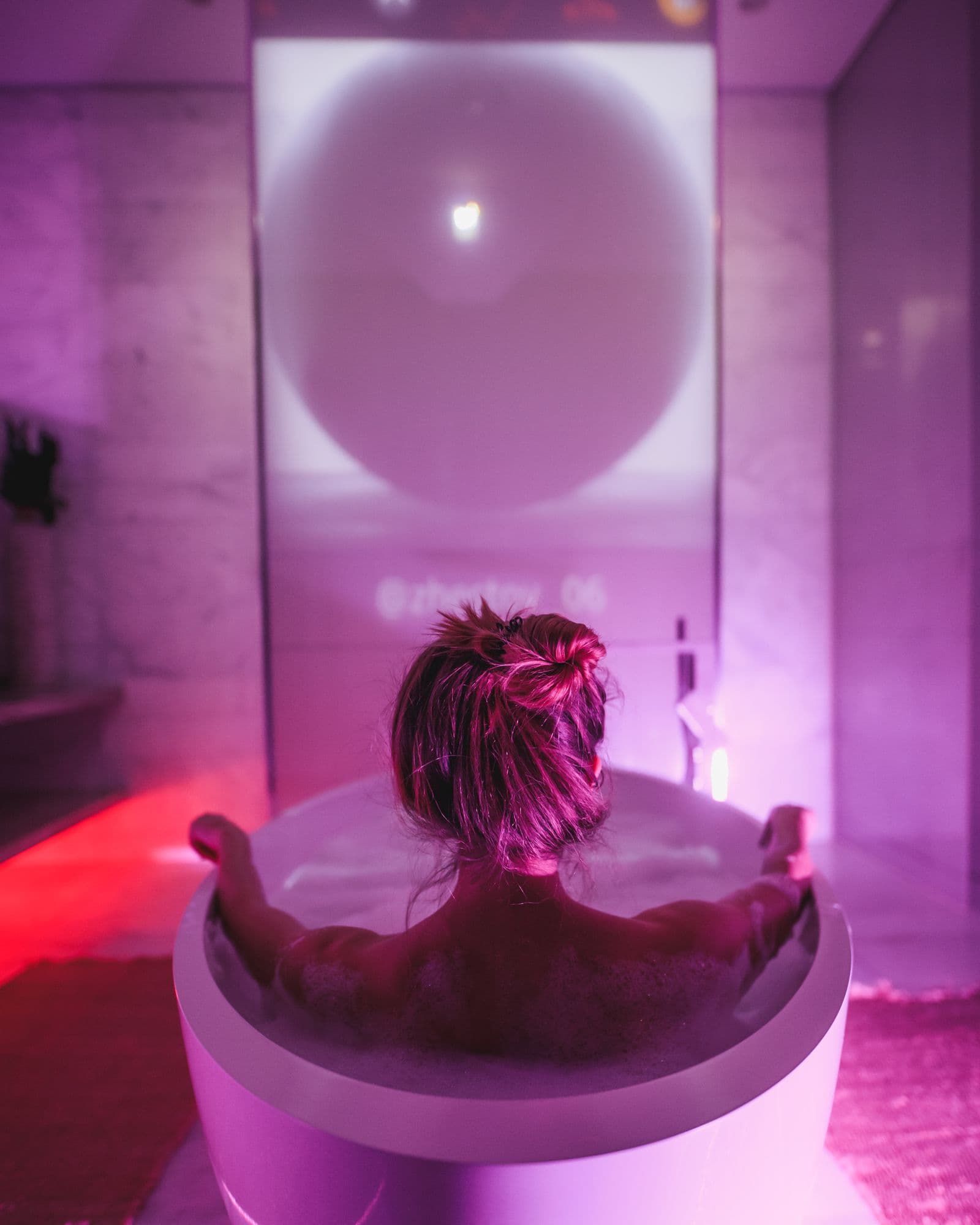 The NFT bath — projection visible on the glass partition reading @klarens, soft pink light through the room.