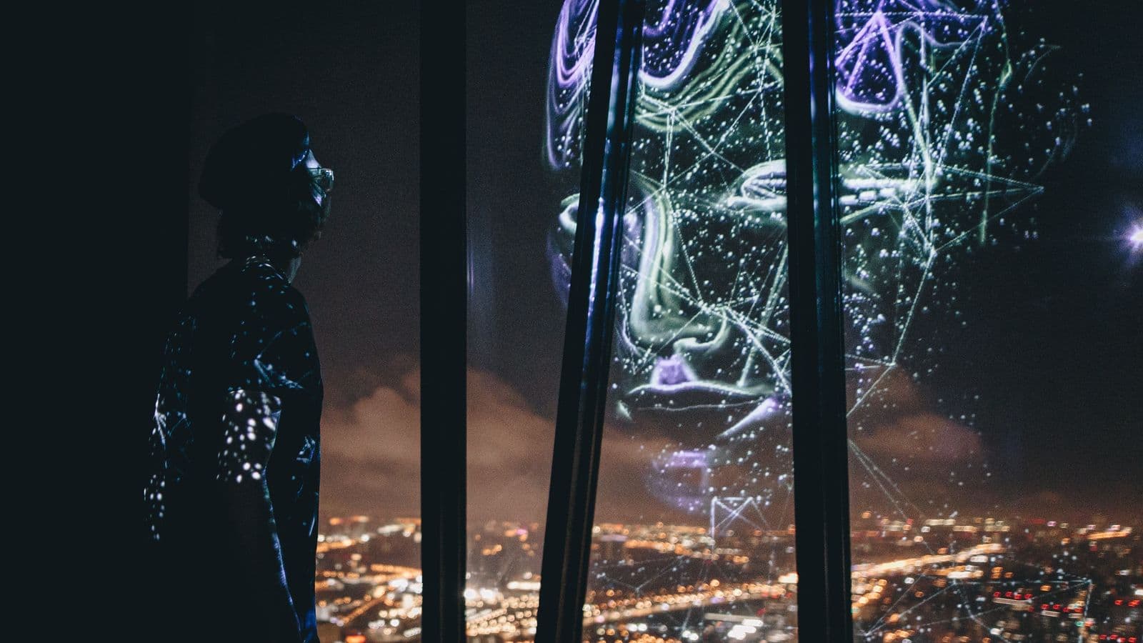 A digital face hologram resolves on the panoramic city window at night.