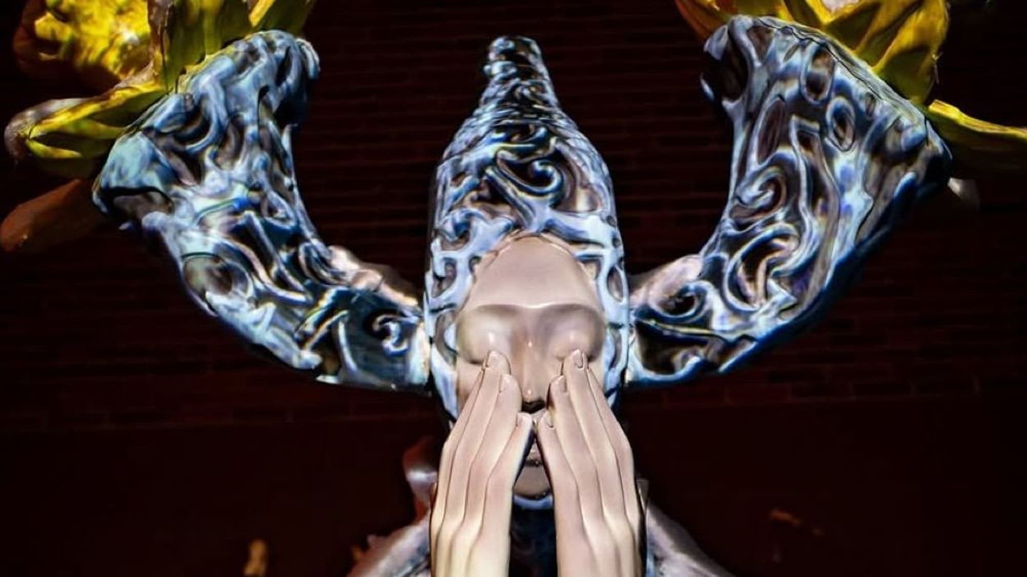 Final state — Ephemera lit at night, hands clasped at the face, baroque blue projection across the robe, golden flowers on the antler crown. Photo by Tyreek B Hitt.